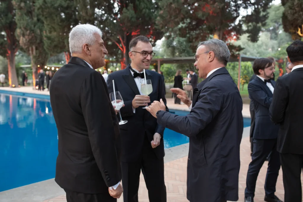 Three men in black tuxedos converse beside a pool at an outdoor formal event, each holding a drink with a straw.