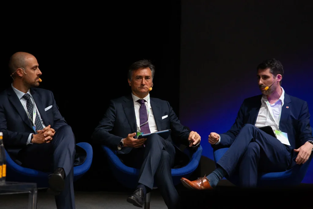 Three men in dark suits seated on a panel stage, each with a headset microphone, engaged in a discussion under stage lighting.